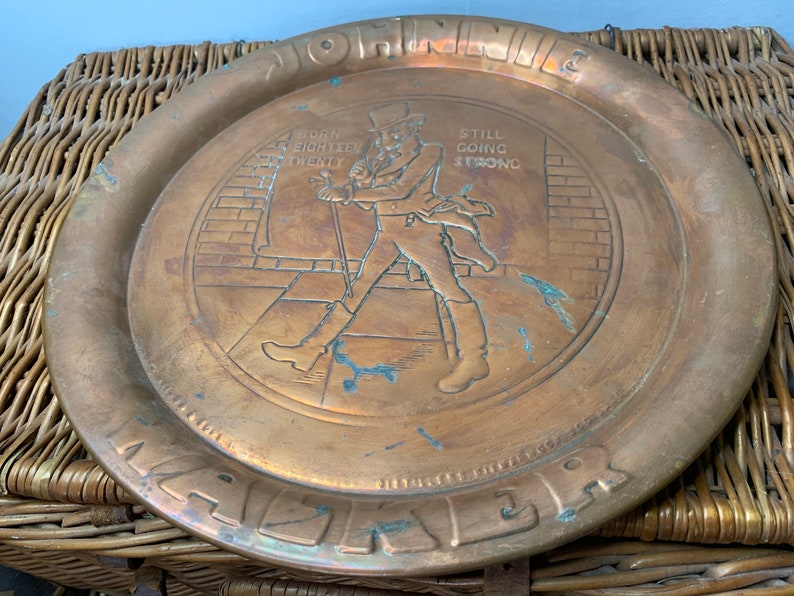 Puede incluir: Una bandeja redonda de lat&oacute;n con una imagen en relieve de un hombre caminando. La bandeja tiene las palabras "Johnnie Walker" y "Born 1820 Still Going Strong" alrededor del borde.