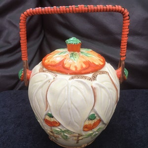 May include: A white ceramic teapot with a lid and a handle. The teapot is decorated with orange and green flowers and leaves. The handle is made of woven orange and black material.