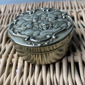 Puede incluir: Caja de alhajas de plata con un diseño en relieve de cuatro ángeles con alas. La caja es redonda y tiene una superficie lisa y pulida.