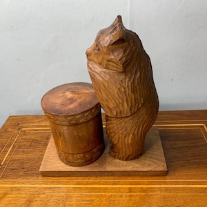 May include: A wooden sculpture of a bear standing on a wooden base with a carved wooden cylinder. The bear is carved in a realistic style with detailed fur and facial features.