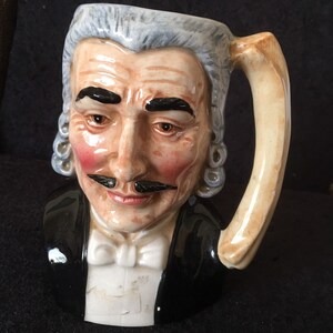 May include: A ceramic mug shaped like a man's bust. The mug is painted to look like a man wearing a black suit with a white bow tie and a white wig. The man has a mustache and a serious expression.