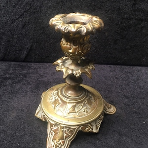 May include: A gold-toned metal candlestick with an ornate design. The base has a three-pronged design with intricate details. The candlestick is approximately 6 inches tall.
