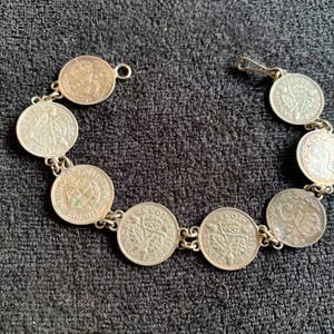 May include: Silver coin bracelet featuring multiple round coins linked together. The coins have various designs and dates, connected by small silver links. The bracelet is displayed on a dark gray textured surface.