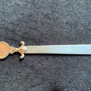 Könnte beinhalten: Silberner Brieföffner mit einem dekorativen Griff, der ein kreisförmiges Design mit einem Wappen zeigt. Die Klinge ist lang und spitz.