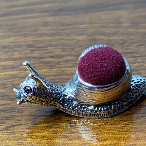 Puede incluir: Un alfiletero de caracol de plata con un cojín de terciopelo burdeos. El caracol tiene una concha texturizada y un solo ojo de piedra preciosa rosa.
