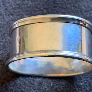 Könnte beinhalten: Ein silberner Serviettenring mit poliertem Finish. Der Ring ist zylindrisch mit abgerundeten Kanten und einer reflektierenden Oberfläche. Das Metall weist einige kleinere Kratzer auf. Geeignet für Tischdekoration und Wohnkultur.