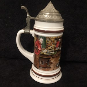 May include: A white ceramic beer stein with a detailed illustration of monks in a library. The stein has a metal hinged lid and handle. The base and top rim are brown.