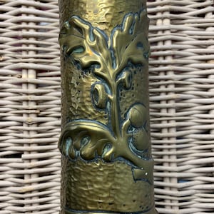 May include: A cylindrical brass vase with a hammered texture and an embossed oak leaf and acorn design. The vase has a slightly flared base and a polished finish, showcasing the intricate details of the botanical motif.