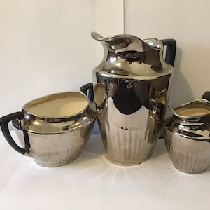 Puede incluir: Un juego de té plateado con una jarra, una jarrita de crema y un azucarero. El juego tiene un acabado brillante y reflectante y un diseño acanalado.