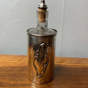 May include: Silver bottle with a hammered finish and a decorative floral design. The bottle has a clear glass body and a cork stopper.