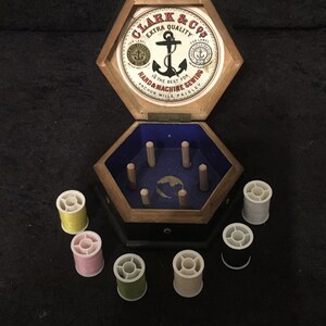 Könnte beinhalten: Eine sechseckige Holzkiste mit blauem Innenraum und einem Deckel mit einem roten und weißen Etikett mit dem Text "CLARK & Co's EXTRA QUALITY IS THE BEST FOR HAND & MACHINE SEWING ANCHOR MILLS. PAISLEY." Die Box enthält sechs Garnrollen in verschiedenen Farben, darunter Gelb, Rosa, Grün und Weiß.
