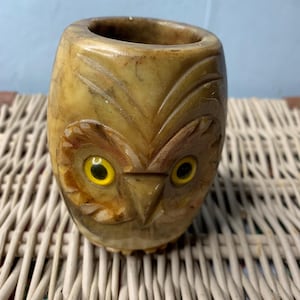 Puede incluir: Un recipiente tallado en forma de búho con ojos amarillos. El recipiente está hecho de una piedra de color marrón claro y tiene forma cilíndrica. La cara del búho está intrincadamente tallada con detalles de plumas y un pico.