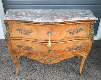 May include: A vintage wooden chest of drawers with a marble top. The chest features curved drawers, ornate brass hardware, and decorative accents. The wood has a warm, natural finish, and the marble top has a gray and red pattern.