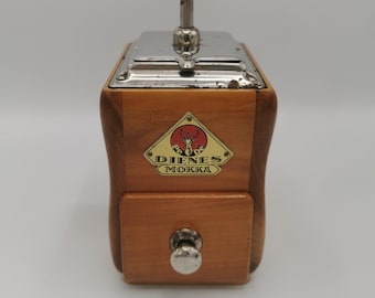 Könnte beinhalten: Eine antike Kaffeemühle aus Holz mit silbernem Oberteil und schwarzem Knopf. Die Mühle hat ein rotes und weißes Etikett mit der Aufschrift "Dienes Mokka" und einem Hirschlogo.