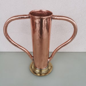 Könnte beinhalten: Eine Kupfervase mit zwei Griffen und einem Messingfuß. Die Vase hat eine gehämmerte Oberfläche und eine zylindrische Form.