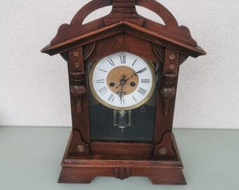 May include: A vintage wooden mantle clock with a Roman numeral clock face and a pendulum. The clock has a decorative, ornate design with a pointed top.