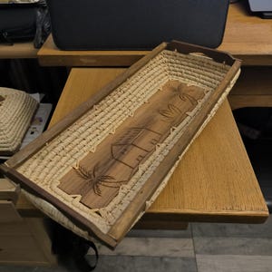 May include: A rectangular woven basket with a wooden frame. The basket has a carved design of a house and palm trees on the bottom.