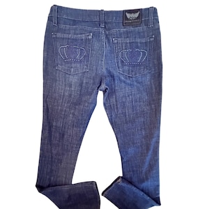 May include: Dark wash denim jeans with a classic five-pocket design. The back pockets feature a crown design with rhinestone accents. A leather patch with the brand logo is on the back waistband.