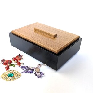 May include: A rectangular wooden box with a light brown lid and a black base. Three decorative brooches are in the foreground: a red bird brooch, a purple hummingbird brooch, and a gold brooch with a green center stone.