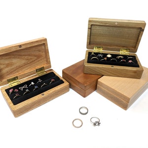 May include: Two wooden ring boxes with black velvet interiors. The boxes are open and filled with rings. The boxes are made of light wood with a natural grain.