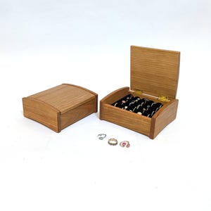 May include: Two wooden jewelry boxes, one closed and one open, displaying rings. The open box reveals a black velvet interior with multiple ring slots. The boxes are made of light-colored wood with a curved lid design.