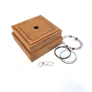 May include: A wooden jewelry box with a decorative etched design on the lid, next to a collection of jewelry. The jewelry includes silver rings, silver and black bangles, and a beaded bracelet with purple and clear beads.