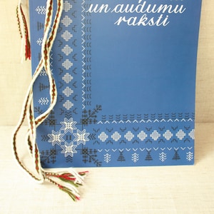 May include: A blue book with the title "Izšuvumu, adījumu un audumu raksti" in white lettering. The book is bound with a white, red, and green ribbon.