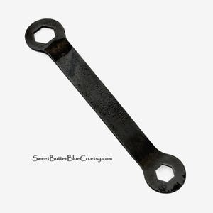 May include: A dark gray metal double-ended wrench with hexagonal openings at both ends. The tool features the stamped text ROCKWELL 1931-X on its flat surface. The metal shows signs of age and wear with a textured, matte finish throughout the body.