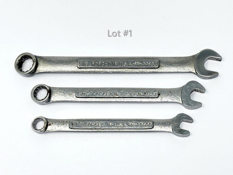 May include: Three silver-colored metal combination wrenches of varying sizes arranged horizontally. Each tool features a box end and an open end, with the text "FORGED IN U.S.A." and specific size markings embossed on the handle. The image includes the text "Lot #1".