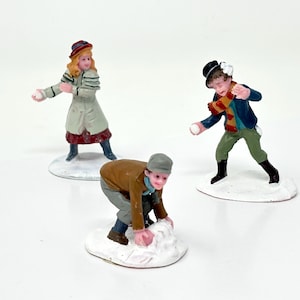 May include: Three miniature figurines depicting a winter scene. A girl in a coat and hat prepares to throw a snowball, while a boy in a scarf and jacket throws one. Another boy is making a snowball. The figurines are on white bases.