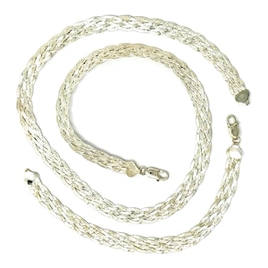 May include: A silver-colored braided necklace with a lobster clasp closure. The necklace is made of multiple strands woven together, creating a flat, wide design. The necklace is displayed on a white background.