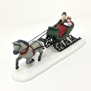 May include: A decorative figurine of a horse-drawn sleigh. The gray horse is pulling a green sleigh with two figures inside. The sleigh is on a white base, suggesting snow. The figures are wearing winter attire.