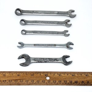May include: A set of five silver-colored metal Craftsman wrenches of varying sizes arranged horizontally above a wooden ruler that measures up to 10 inches. Each wrench features embossed text indicating the brand and size specifications.