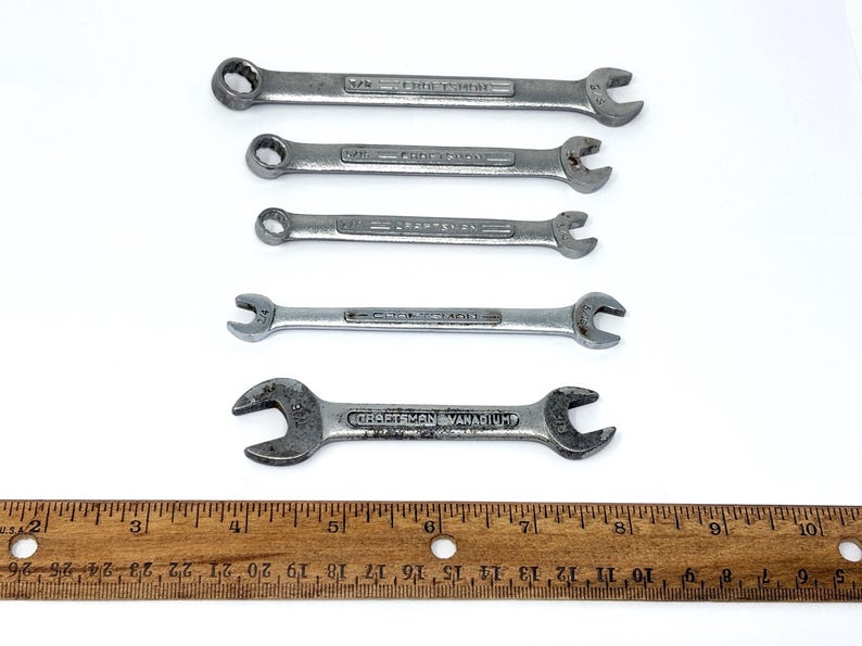 May include: A set of five silver-colored metal Craftsman wrenches of varying sizes arranged horizontally above a wooden ruler that measures up to 10 inches. Each wrench features embossed text indicating the brand and size specifications.