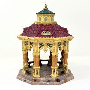 May include: A miniature gazebo with a burgundy tile roof, green top, and ornate gold columns. The gazebo has benches inside and sits on a stone-like base. A decorative piece for a miniature village or display.