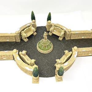 May include: A miniature garden display featuring a central tiered fountain with a statue, surrounded by four stone-colored benches with lion-shaped ends, low stone walls, and four small green conical topiary trees on a dark cobblestone base.