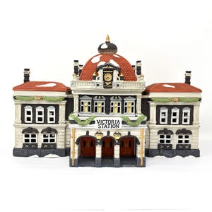 May include: A ceramic Victoria Station Christmas village building with a red and white roof, a clock, and the words "Victoria Station" on the front. The building has multiple windows and doors, and is decorated with green garland and snow.