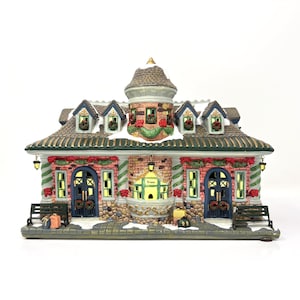 May include: A detailed Christmas village building with a brick facade, arched windows, and a central tower. The building features a snow-covered roof, wreaths, and festive decorations. The word "Tickets" is visible above the entrance.