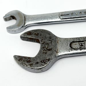 May include: Two silver-colored metal wrenches with open-ended heads. The top wrench has a smooth, polished finish with the text FORGED IN visible. The bottom wrench has a weathered, textured surface with rust spots and the number 1723 stamped on it.