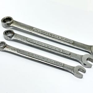 May include: Three silver-colored metal combination wrenches arranged diagonally. Each tool features a box end and an open end, with the text FORGED IN U.S.A. and specific size markings clearly stamped into the metal surface of the handles.