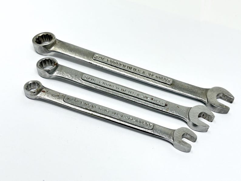 May include: Three silver-colored metal combination wrenches arranged diagonally. Each tool features a box end and an open end, with the text FORGED IN U.S.A. and specific size markings clearly stamped into the metal surface of the handles.
