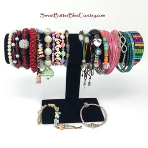 May include: A collection of colorful bracelets on a black display stand. The bracelets are made of various materials, including beads, leather, and metal. Some of the bracelets feature charms, such as a heart, a flower, and an infinity symbol. The text "SweetButterBlueCo.etsy.com" is visible in the top center of the image.