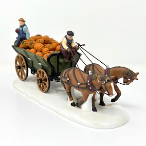 May include: A decorative figurine depicting a harvest scene. A green wagon filled with orange pumpkins is pulled by two brown horses. Two figures are in the wagon. The figurine is set on a white base.