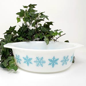 May include: White oval casserole dish with light blue snowflake pattern. The dish has two handles and is surrounded by green foliage. A vintage kitchenware item.