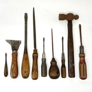 May include: A collection of vintage hand tools, including screwdrivers, a scraper, and a hammer, arranged on a white surface. The tools have wooden handles and show signs of wear, with a rustic, antique aesthetic.