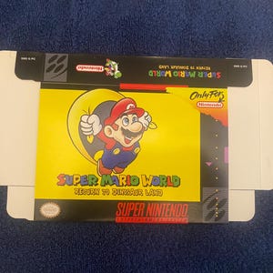 May include: A Super Nintendo game box for Super Mario World, featuring a yellow background with a cartoon image of Mario in a green suit, jumping in the air. The text on the box reads "Super Mario World, Return to Dinosaur Land".