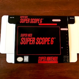 Puede incluir: Una caja de cartón blanca con un cartucho de juego Super Nintendo Super Scope 6 negro y rojo en su interior. La caja tiene el texto "Super NES Super Scope 6" impreso en letras rojas.