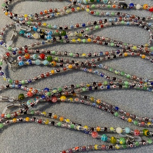 May include: A collection of colorful beaded eyeglass chains. The chains are made with small, round beads in various colors and patterns, including clear, white, black, red, blue, green, and yellow. The chains are strung on a silver cord and have metal clasps at each end.