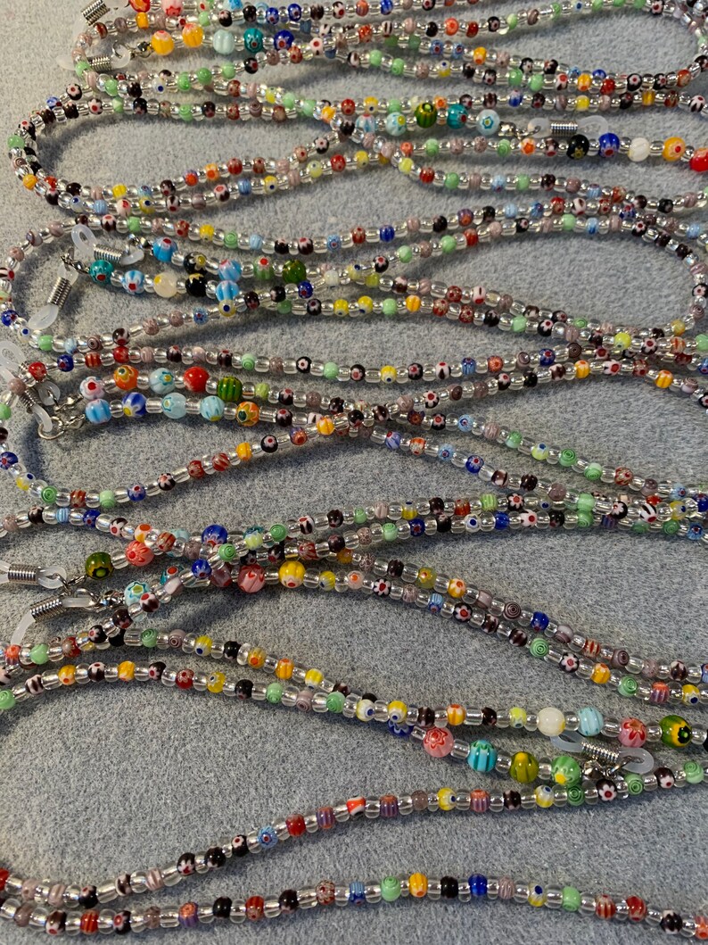 May include: A collection of colorful beaded eyeglass chains. The chains are made with small, round beads in various colors and patterns, including clear, white, black, red, blue, green, and yellow. The chains are strung on a silver cord and have metal clasps at each end.