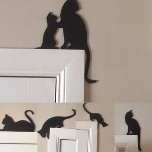 Op de afbeelding: Zwarte silhouetten van katten op een wit deurkozijn. De katten zijn in verschillende poses, waaronder zittend, staand en springend. De silhouetten zijn uit metaal geknipt en zijn met lijm aan het deurkozijn bevestigd.
